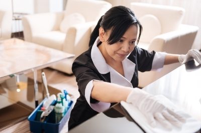 a house maid on hire in disposable gloves, cleaning top shelf of a home